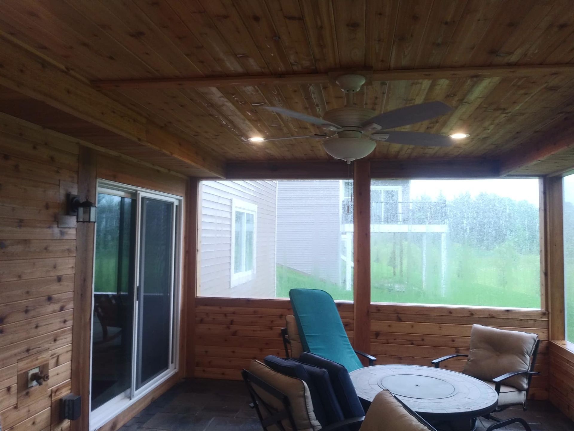Covered patio with cedar walls and ceiling, furnished with chairs and a fire pit, overlooking a rainy yard.