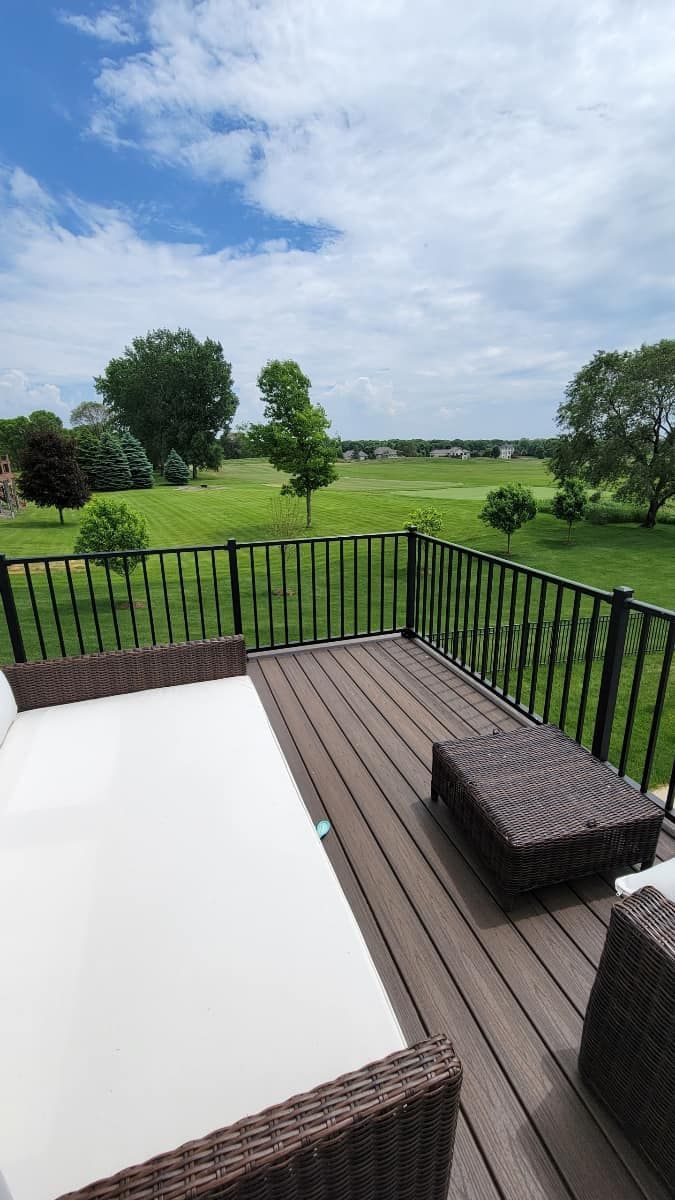 Deck with brown furniture overlooking a green field, trees, and a blue sky with clouds.