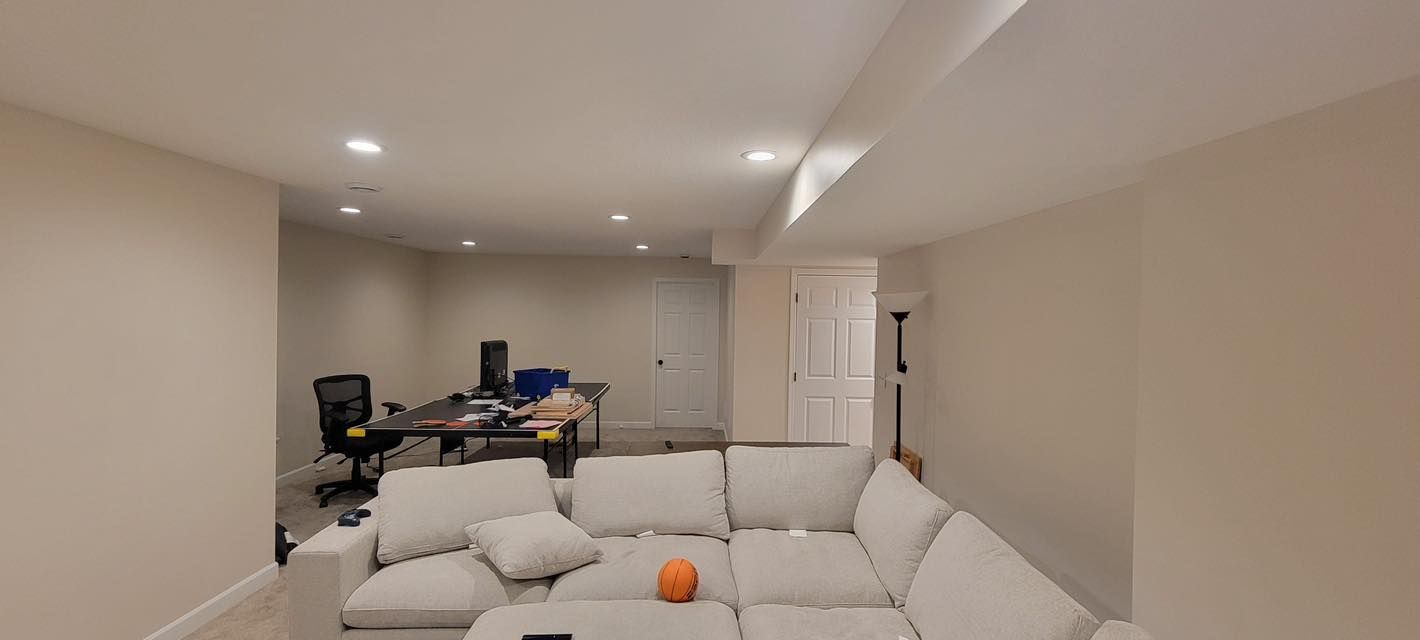 A beige basement living space with a sectional sofa, workspace with a desk and chairs, and a white door.