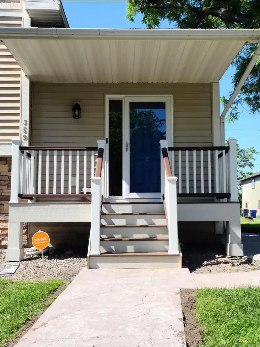 Tan house front porch with steps, railing, and door. Concrete walkway.