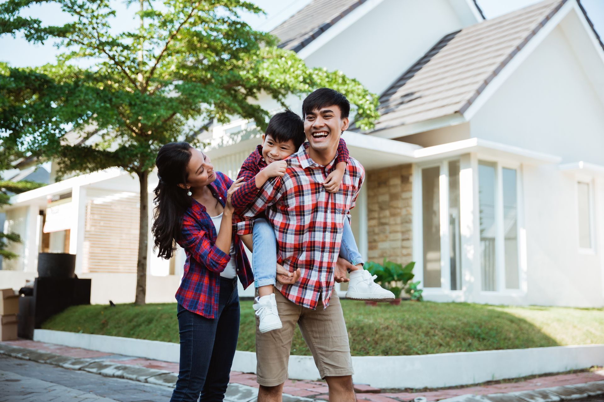 A father giving a piggyback ride to his son while the mother watches in front of a house.