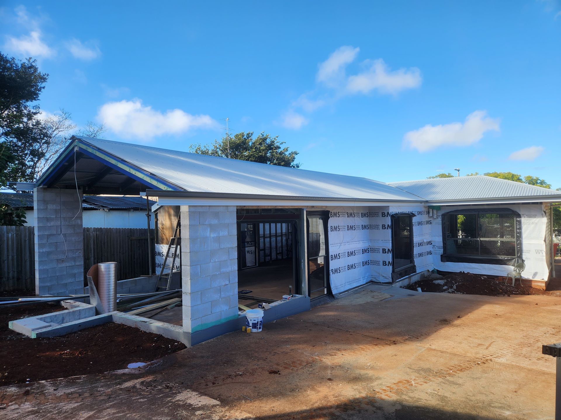 House under construction with concrete block walls, roofing, and tarp-covered windows against a blue sky.