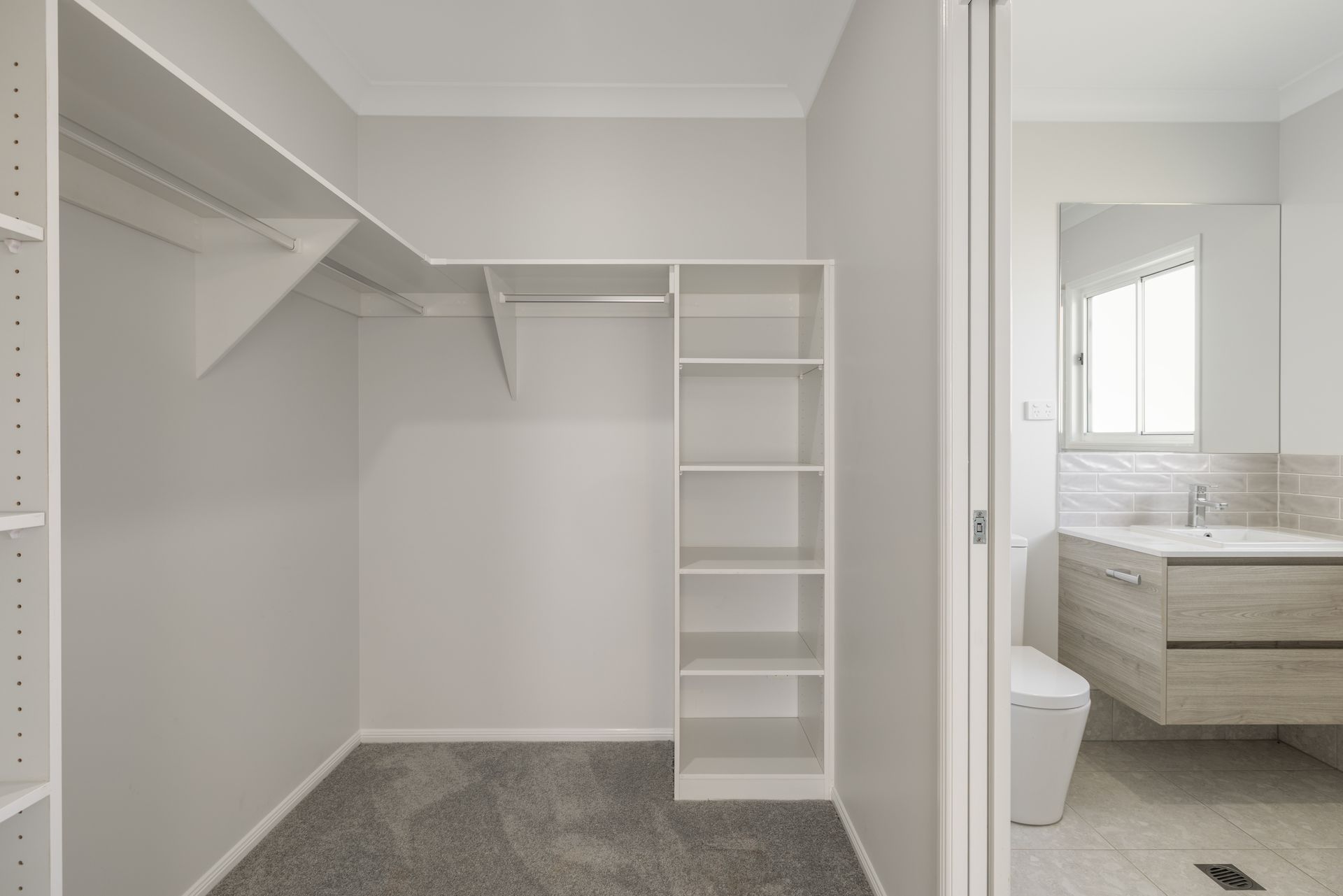 Empty walk-in closet with shelves and a hanging rod, adjacent to a bathroom with a sink and toilet.