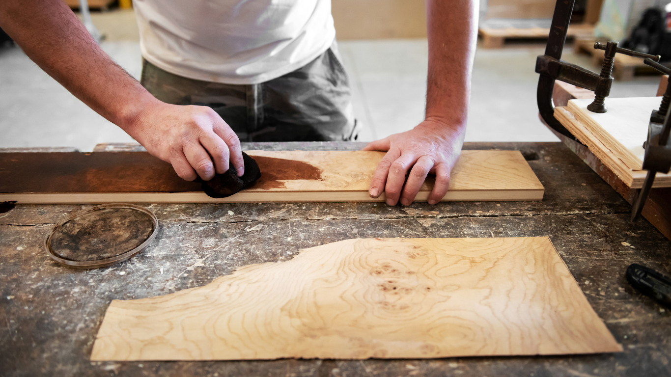Person staining a wooden plank with a cloth in a workshop, another piece of wood in the foreground.