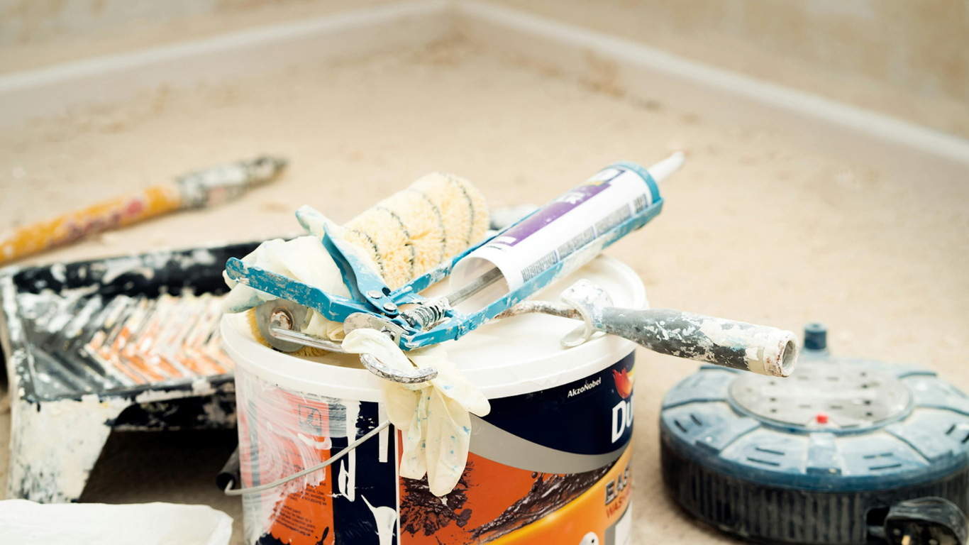 Paint supplies, including a paint can, roller, caulk gun, and paint tray, covered in white paint on a light-colored surface.