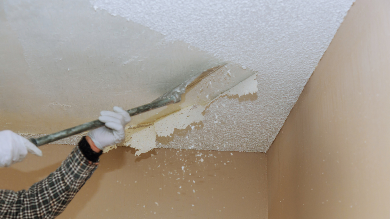 Person removing textured ceiling with a scraper tool. White gloves and arm visible. Beige walls surround.