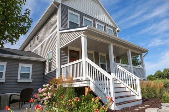 Two-story house with gray siding, white porch, and steps. Landscaping includes flowers and tall grasses under a blue sky.