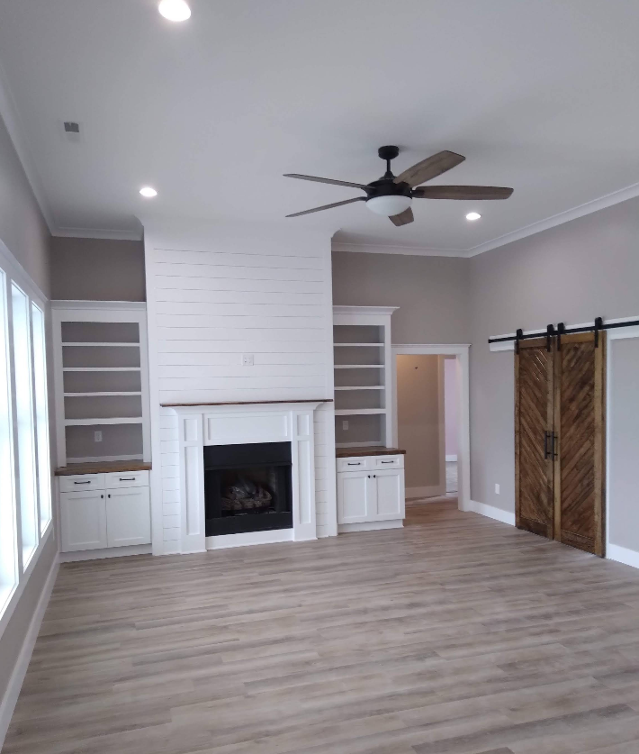 A living room with a fireplace, built-in bookshelves, light wood floors, and a brown door. A ceiling fan is centered above.