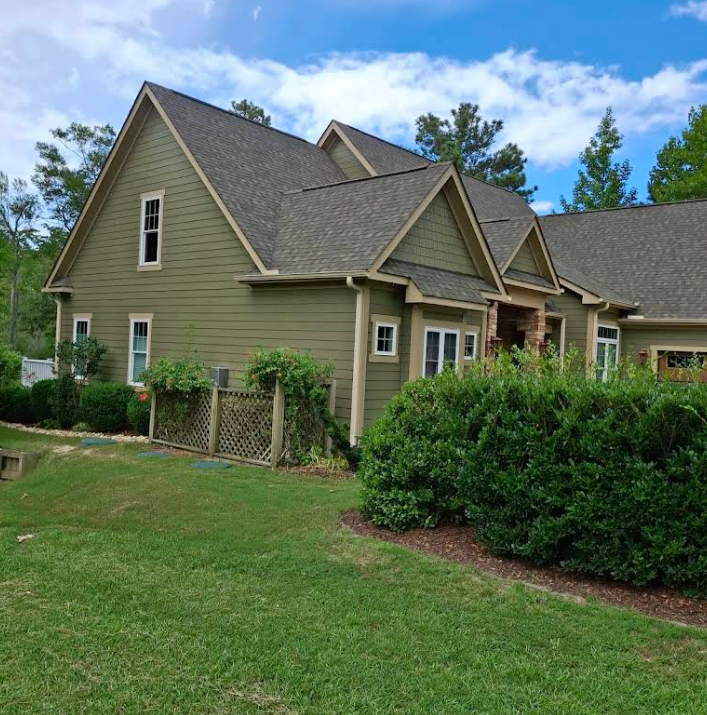 Green house with brown roof and trim, lush green grass and bushes in front.