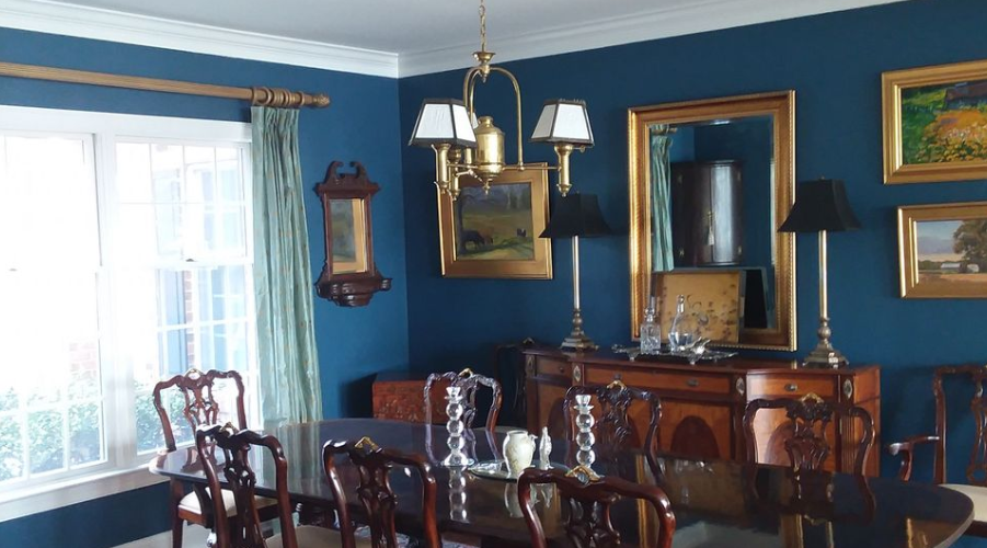 Dining room with a dark blue wall, antique furniture, a chandelier, and framed artwork. A window has pale green drapes.