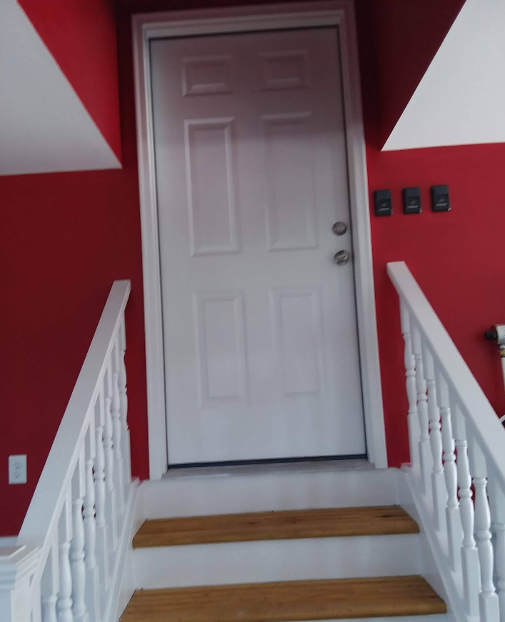 A white door at the top of wooden stairs, with white banisters and red walls. Three black switches at the right of the door.