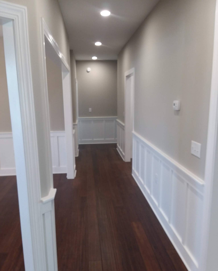 A narrow hallway with dark wooden floors, gray walls, and white wainscoting. Several doorways and ceiling lights are visible.