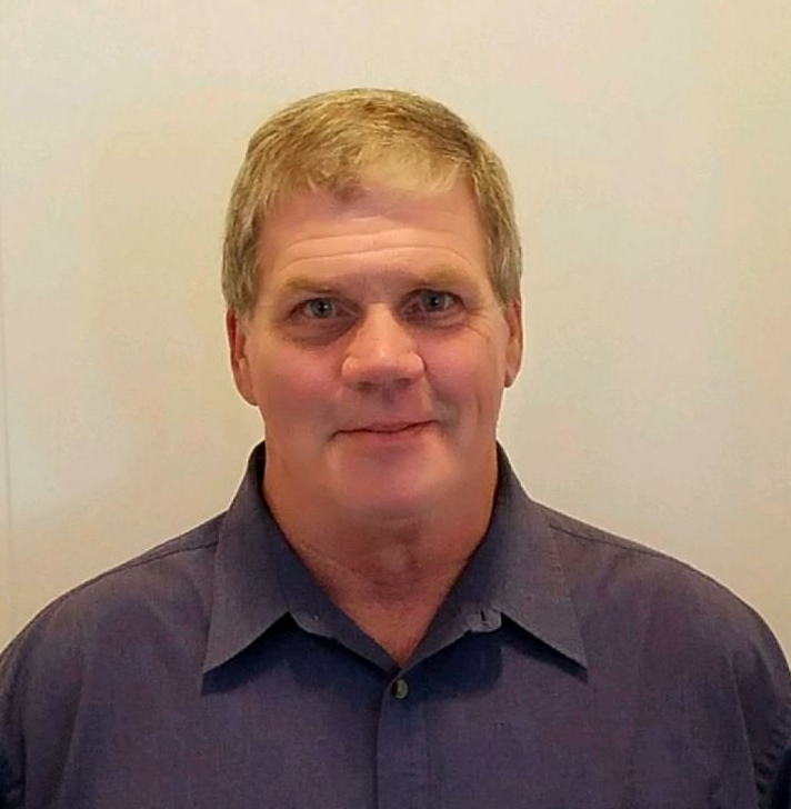 A man with short blond hair, wearing a blue button-down shirt, smiles slightly. He poses in front of a plain wall.