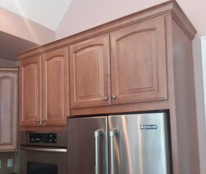 Upper kitchen cabinets with a crown molding above a stainless steel refrigerator and an oven. The cabinets are light brown.