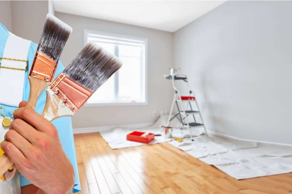 Person holding paintbrushes in a room being painted, with a ladder and drop cloth visible.