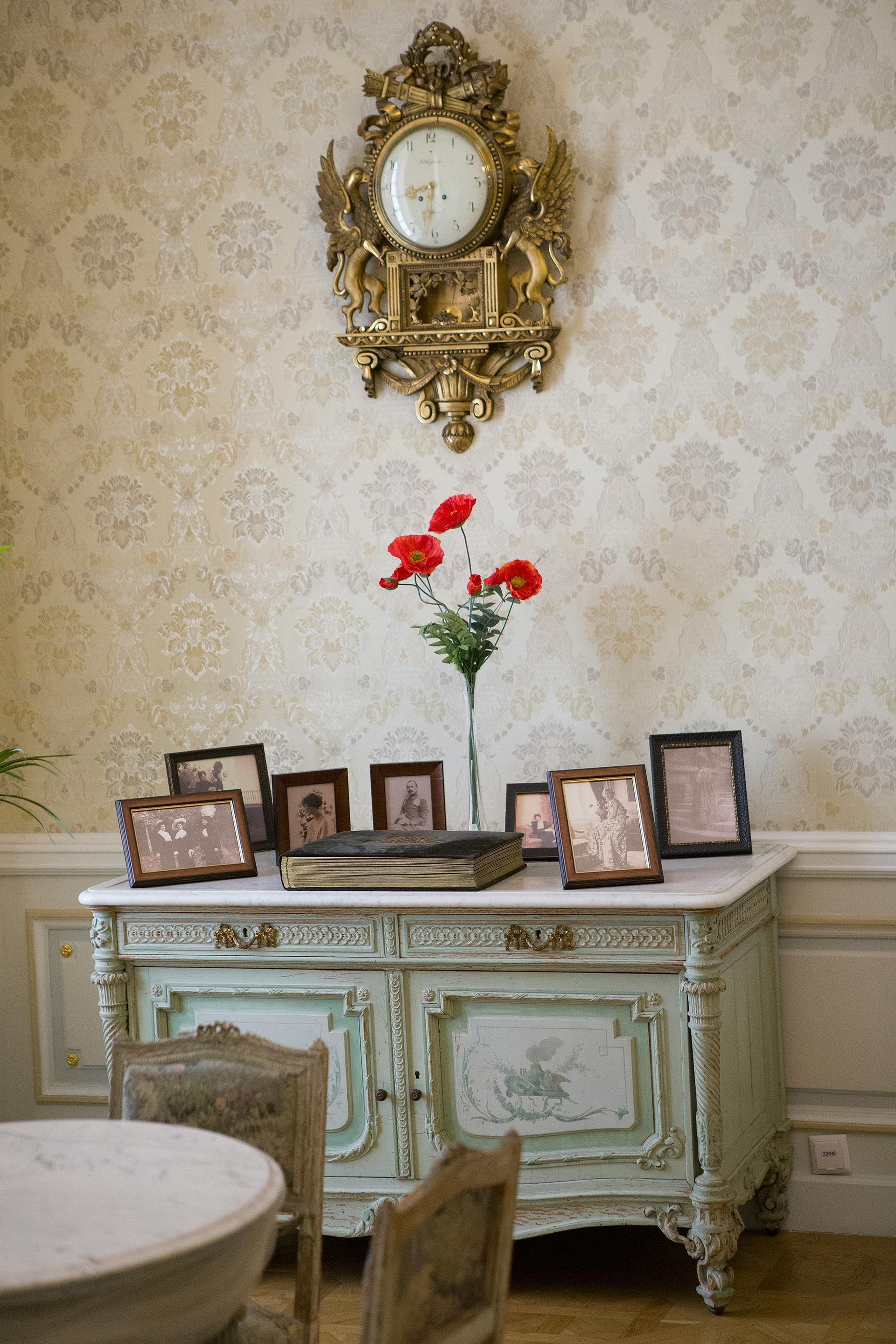 Ornate antique cabinet beneath gilded wall clock, with framed photos and red flowers in a vintage room