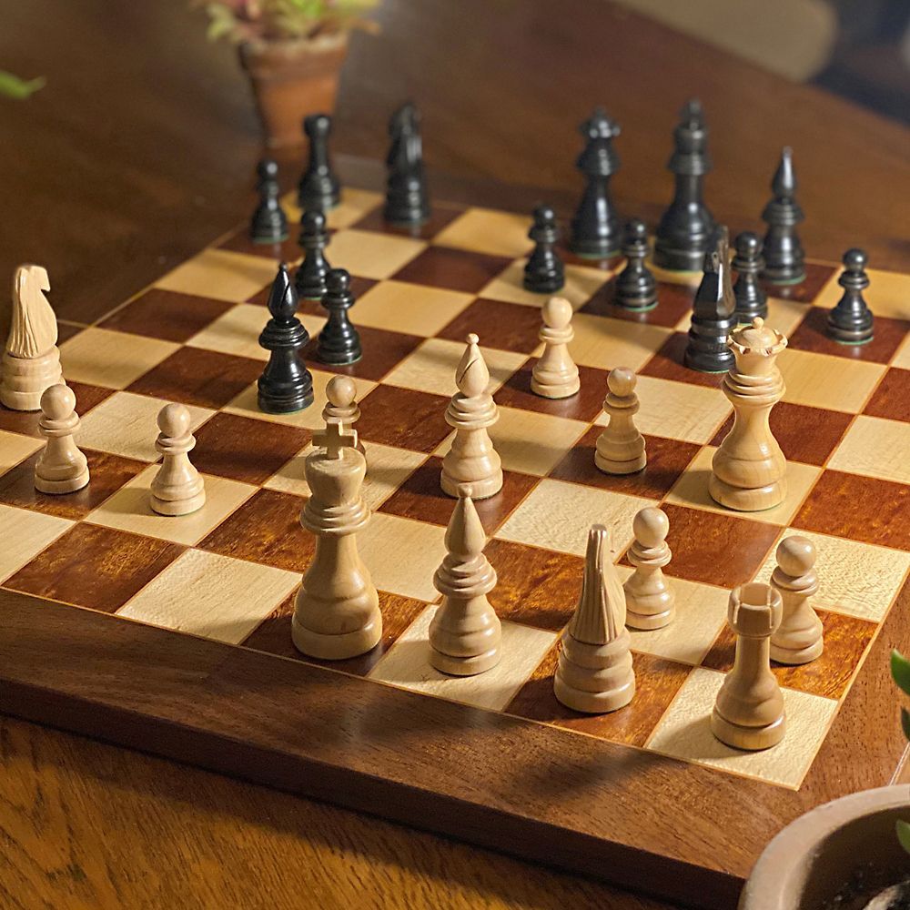 A brown and cream colored wooden chess board with black and cream colored chess pieces on it.