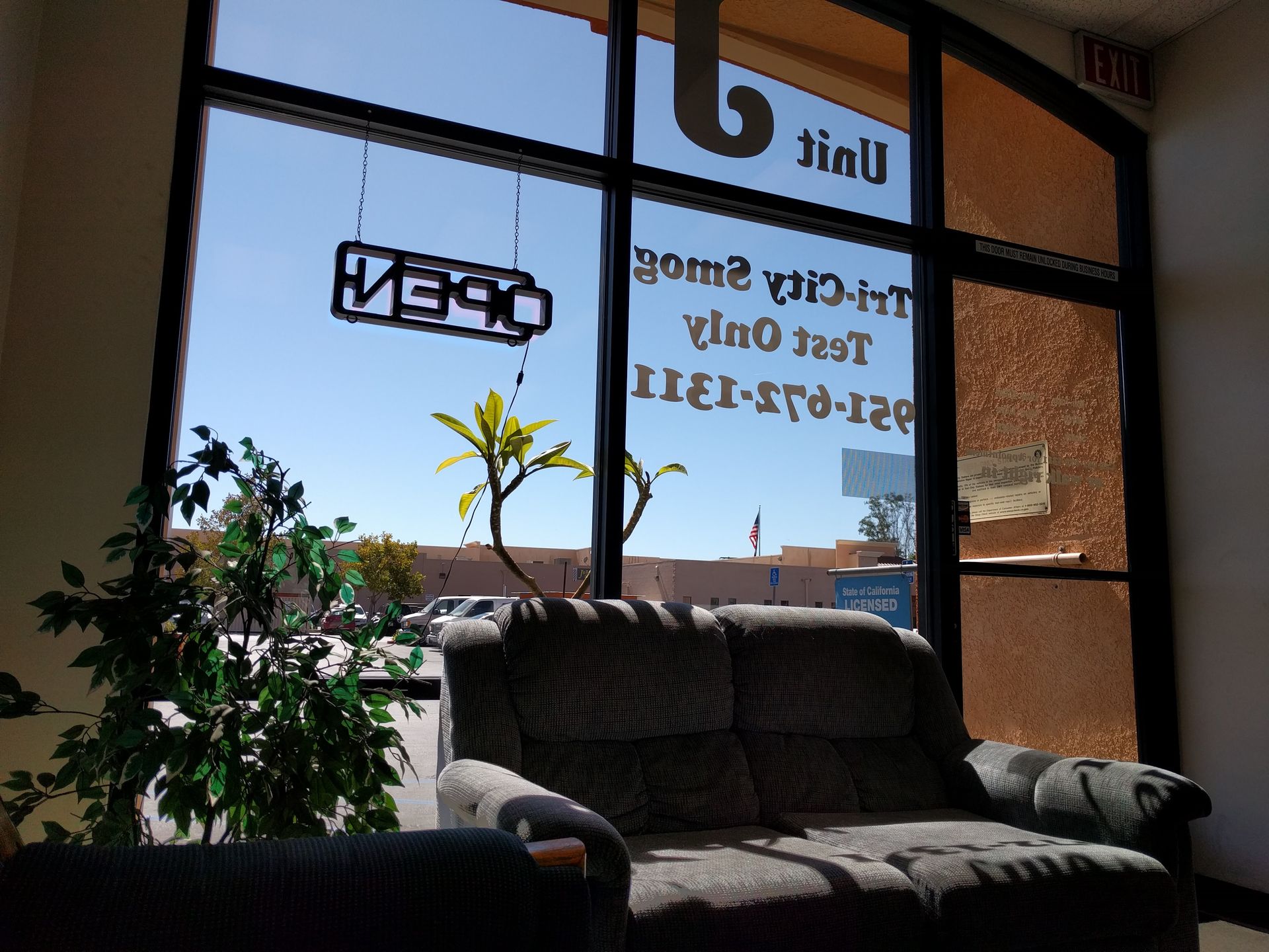Interior view: waiting area with a sofa, plant, and open sign in front of a window. Clear blue sky outside.