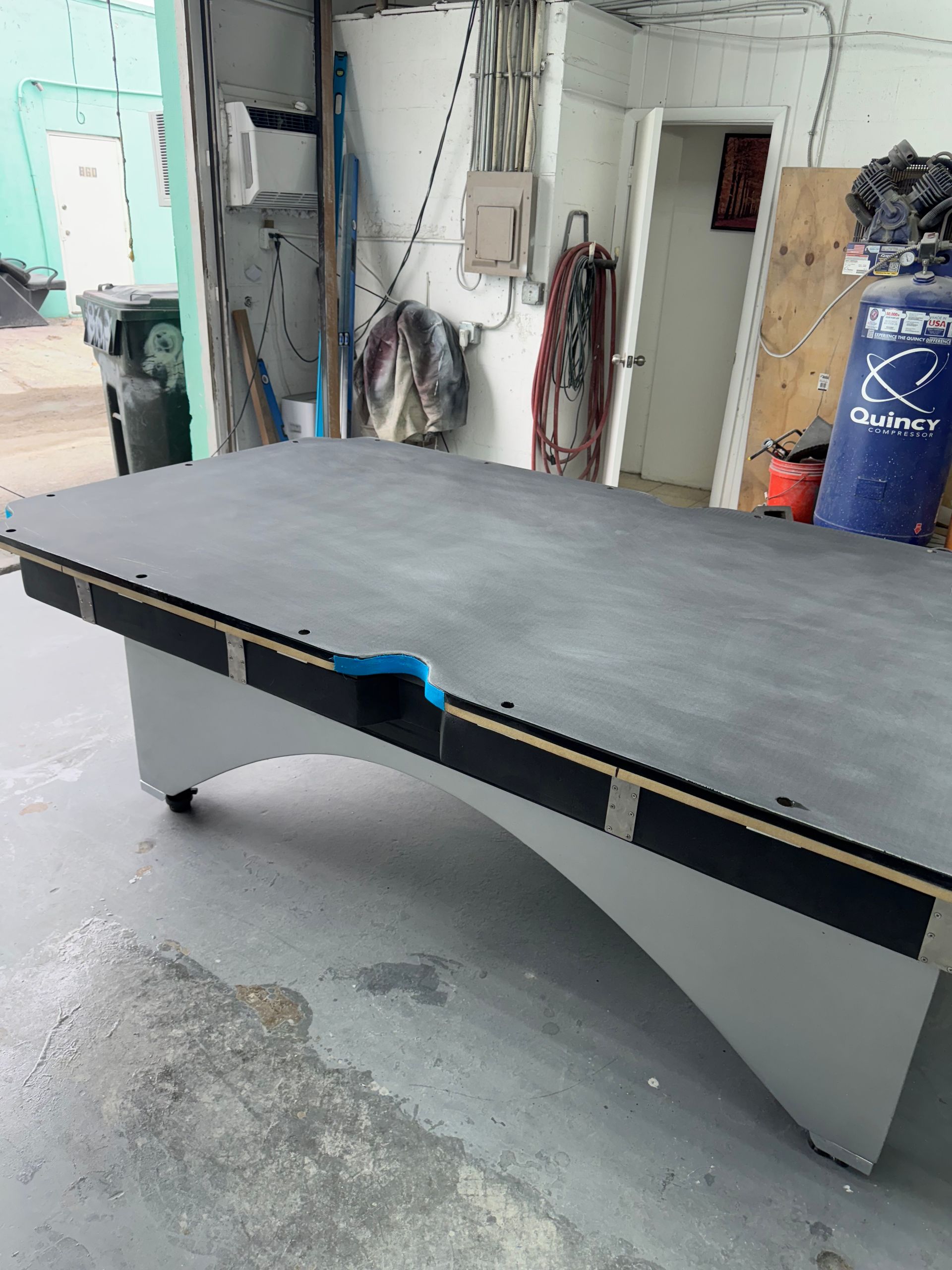 Pool table with gray top and legs, black bumpers, blue pocket, and a workshop backdrop.