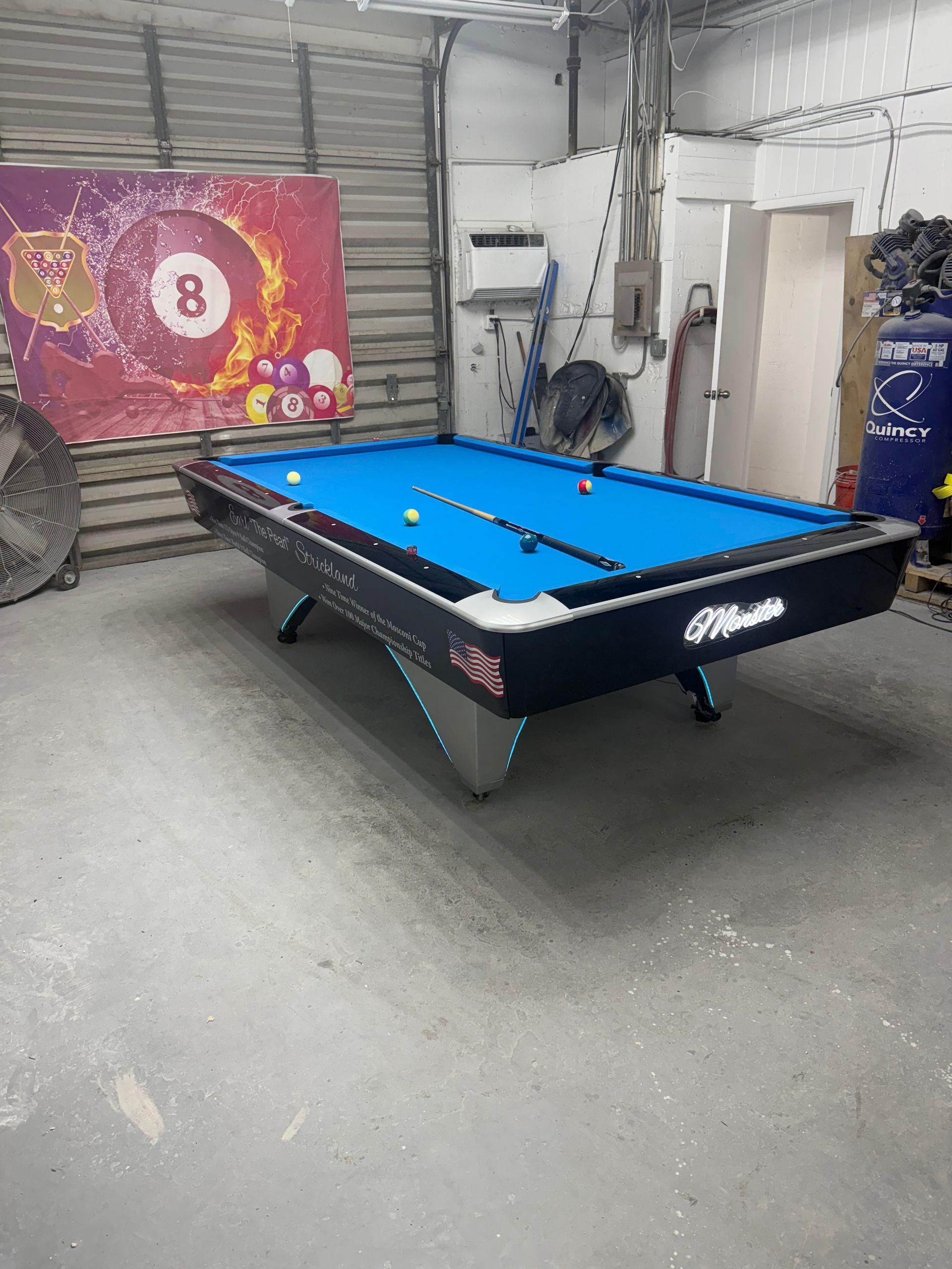 Pool table in a garage with blue felt, balls, and a cue. An 8-ball poster is visible on the wall.