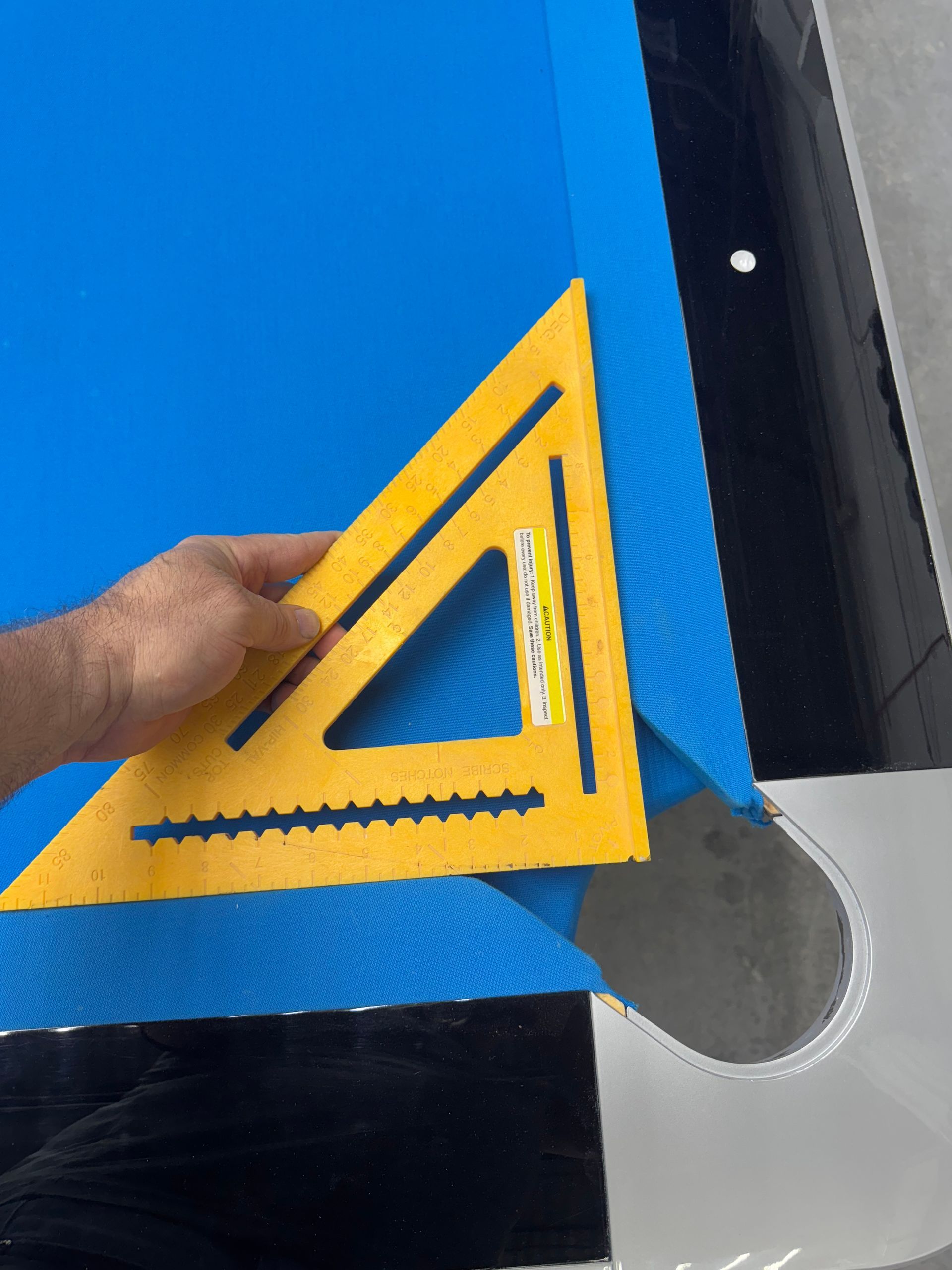 Person using a yellow speed square to check a corner of blue billiard table felt.