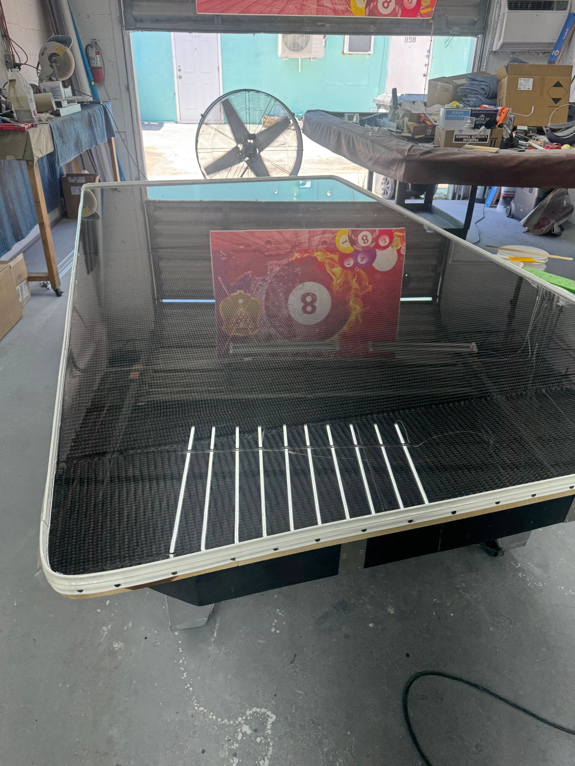 A rectangular screen printing table with a black mesh surface, an 8-ball box is in the center, a fan in the background.