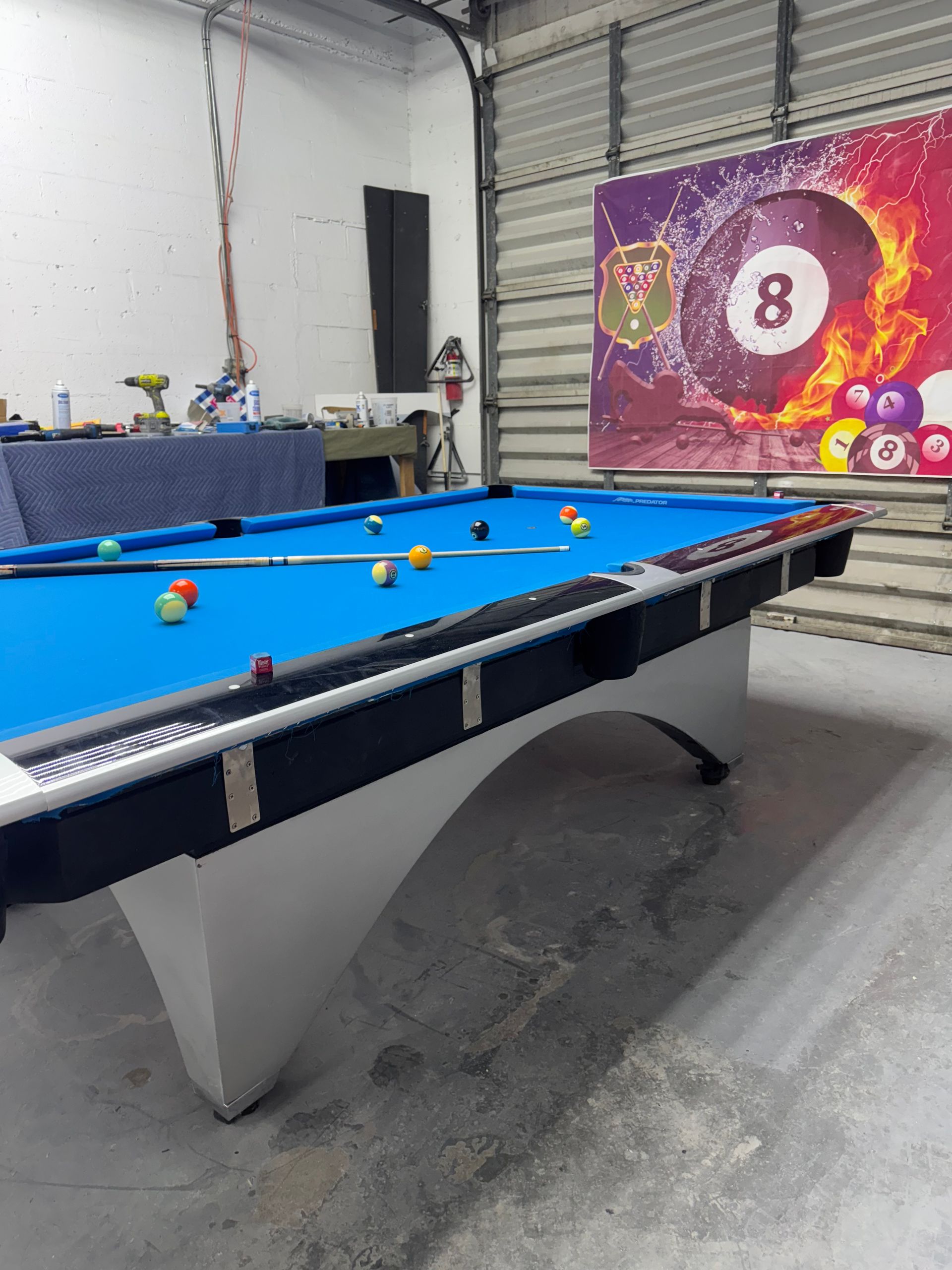 Pool table with blue felt and balls set up, in a garage with an 8-ball graphic on the wall.