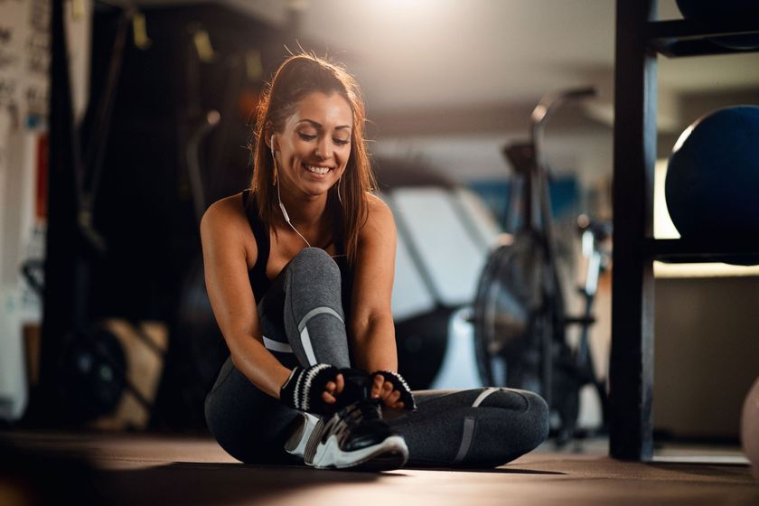Woman in workout clothes tying her shoe in a gym, smiling.