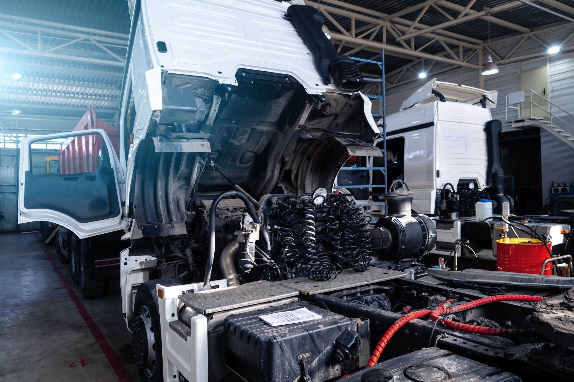 A white semi-truck with its cab tilted forward for engine maintenance inside a well-lit workshop.