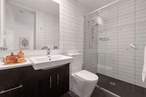 Modern white-tiled bathroom with dark vanity, sink, toilet, and glass shower stall