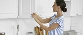 Woman in blue jeans and striped shirt reaching into white kitchen cabinet.
