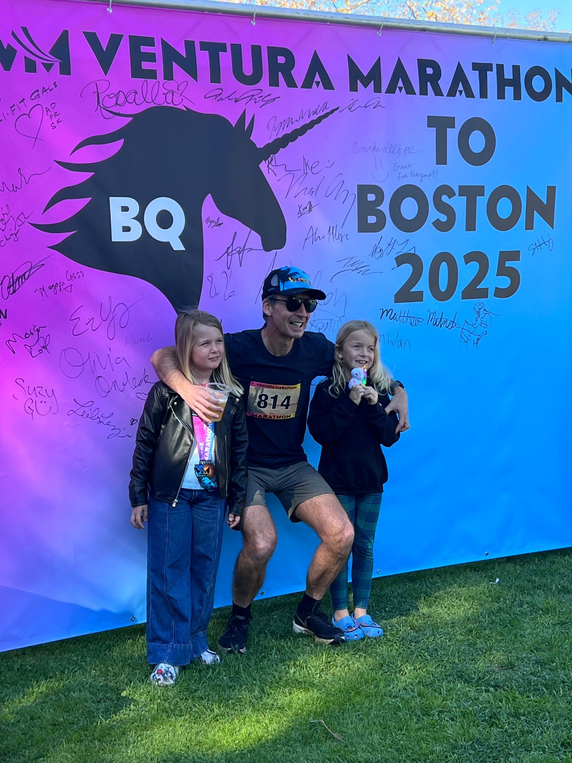 Runner with two kids standing in front of the Boston 2025 banner which has signatures from runners who qualified for the Boston Marathon at the 2025 Ventura Marathon.
