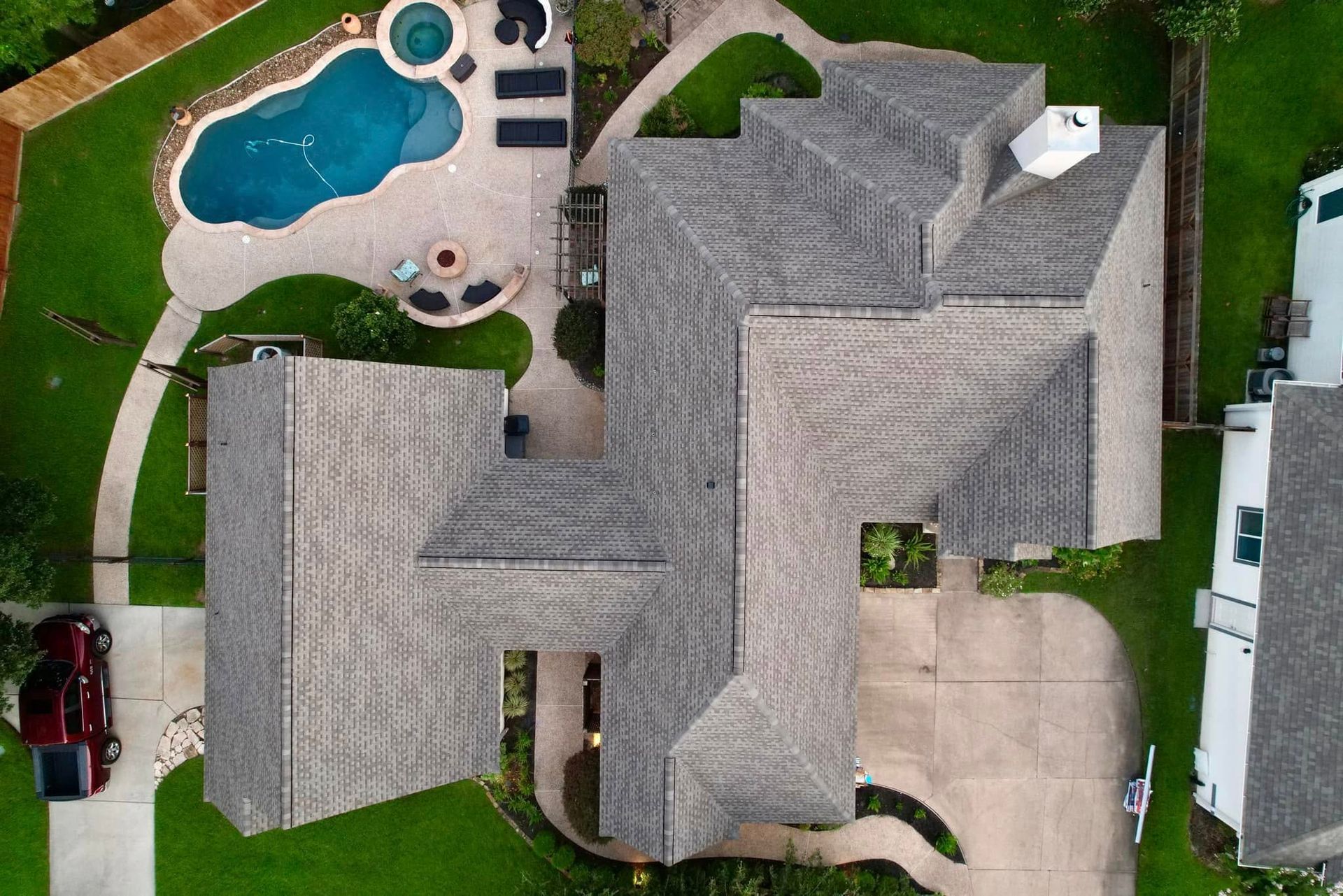 Aerial view of a residential house with a complex grey shingled roof, a backyard pool, and a paved driveway.