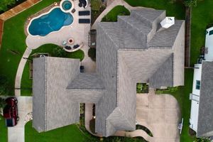 An aerial view of a gray-roofed house with a backyard swimming pool, patio area, and driveway surrounded by green lawns.