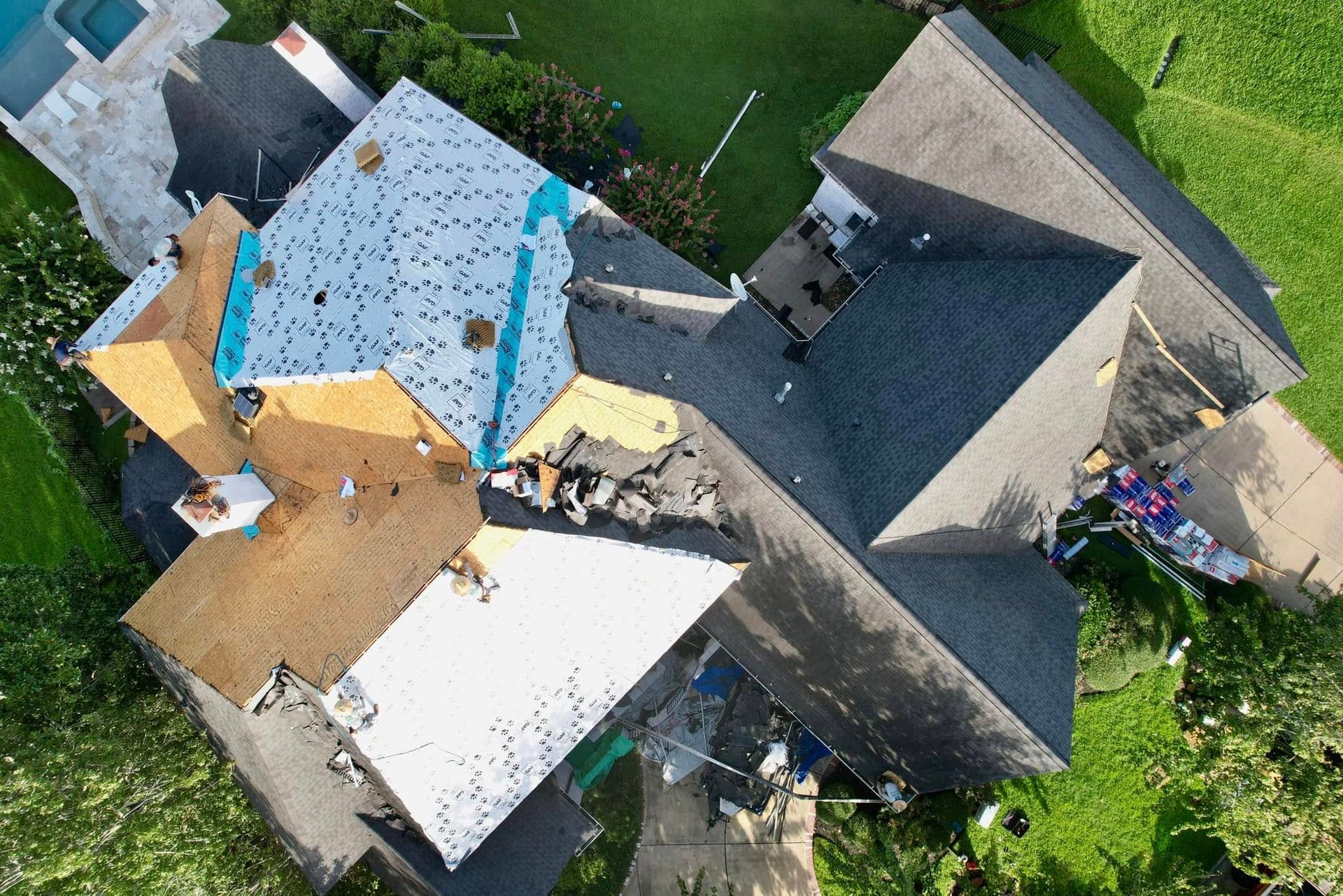 Aerial view of a multi-level residential roof under construction, showing patches of bare wood, underlayment, and shingles.