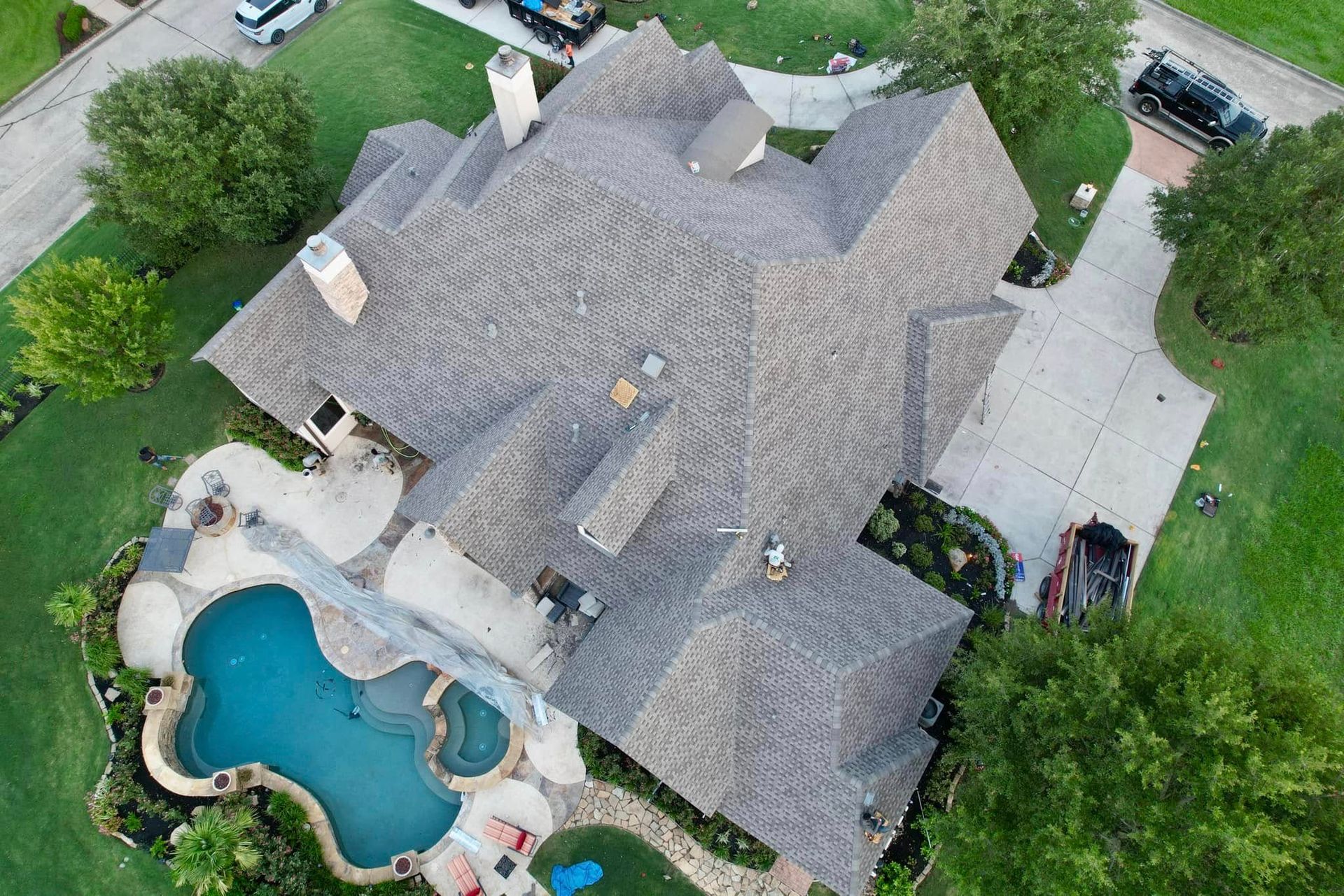 Aerial view of a large residential home with a complex shingled roof, a swimming pool, and a concrete driveway.