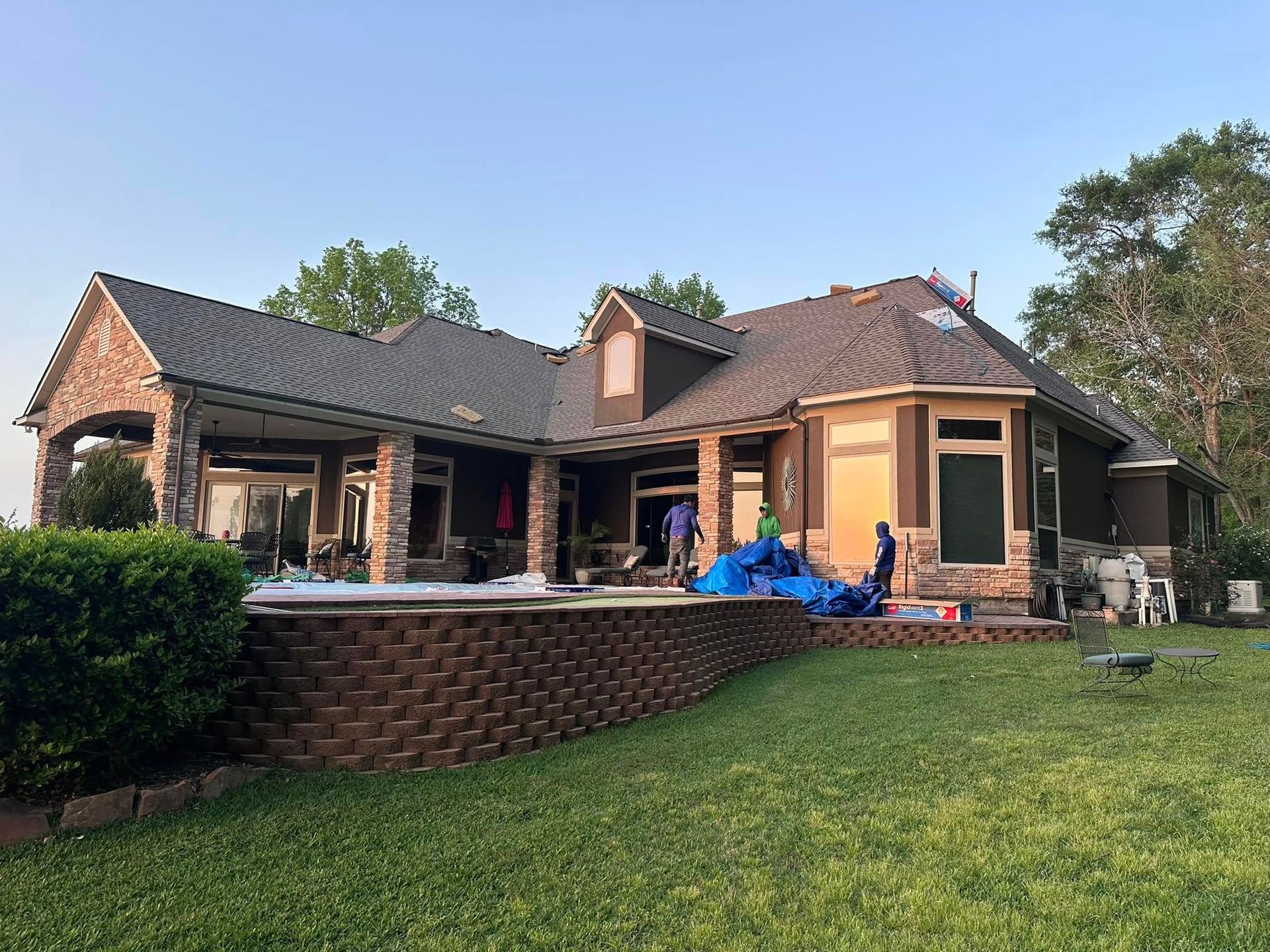 Workers stand on the covered patio of a brown house with stone columns, overlooking a yard and a brick-walled pool area.