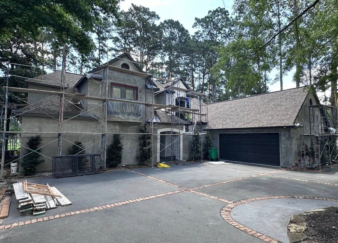 A large two-story house under renovation, surrounded by scaffolding, with a dark garage door and a paved driveway.