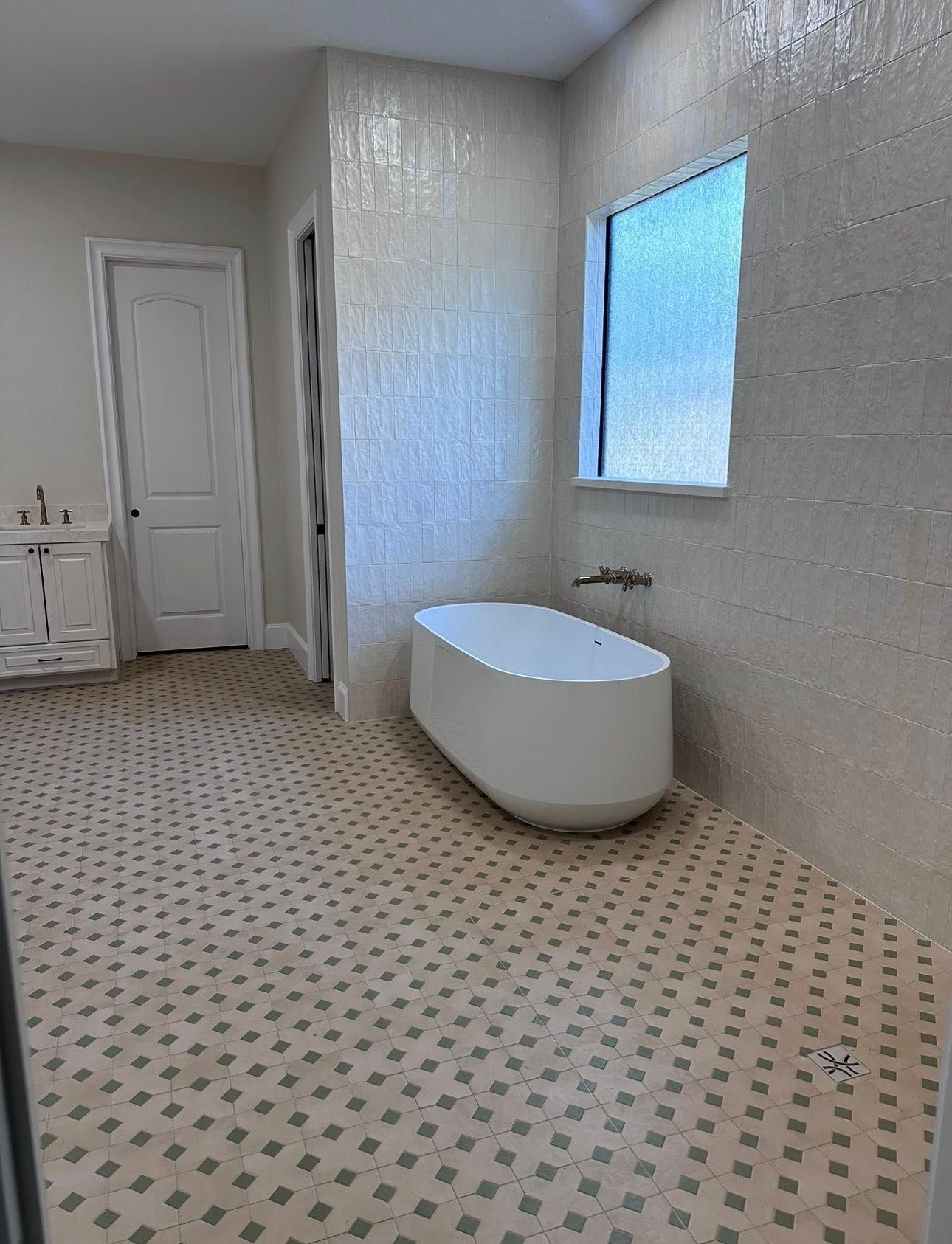 A modern bathroom with a freestanding white bathtub, textured cream wall tiles, patterned tiled floor, and a window.