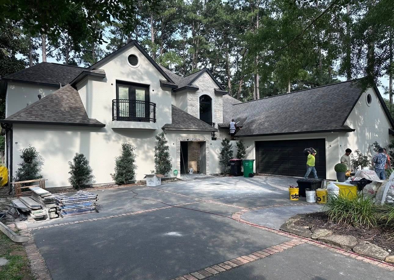 A two-story white house with a dark roof and garage undergoing exterior work, with workers present in the driveway.