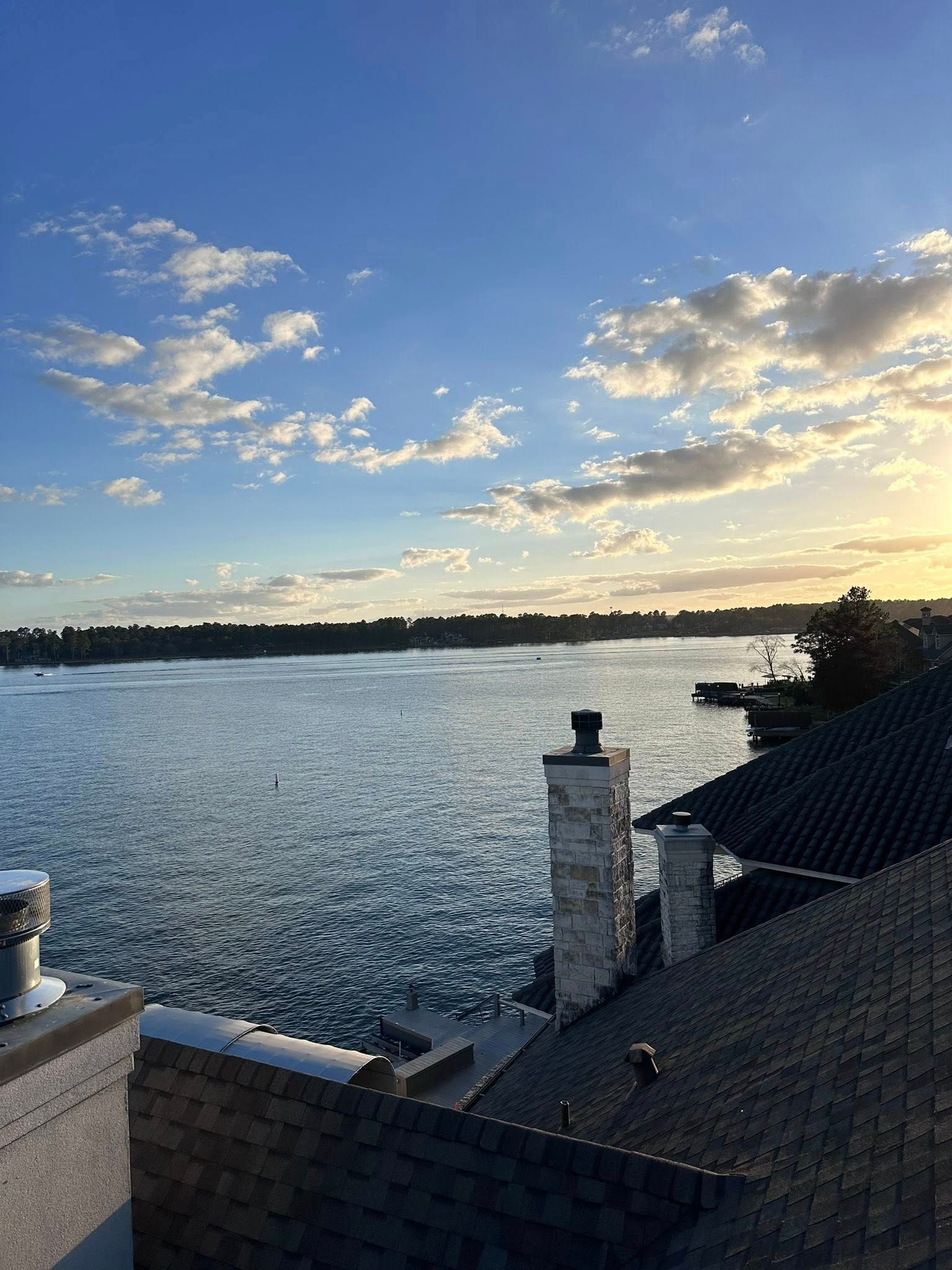 A sunset view over a calm lake, seen from a rooftop with chimneys and dark shingles in the foreground.