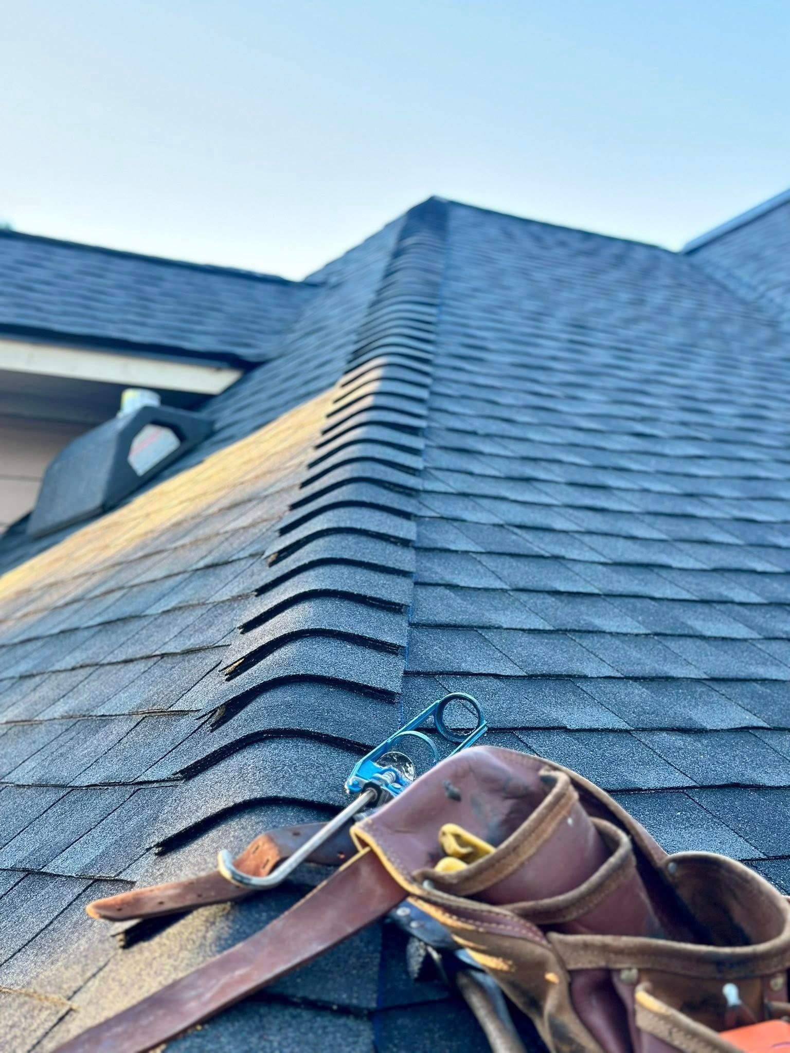 A brown tool belt rests on a steep, dark shingled roof with a partial section of exposed underlayment.