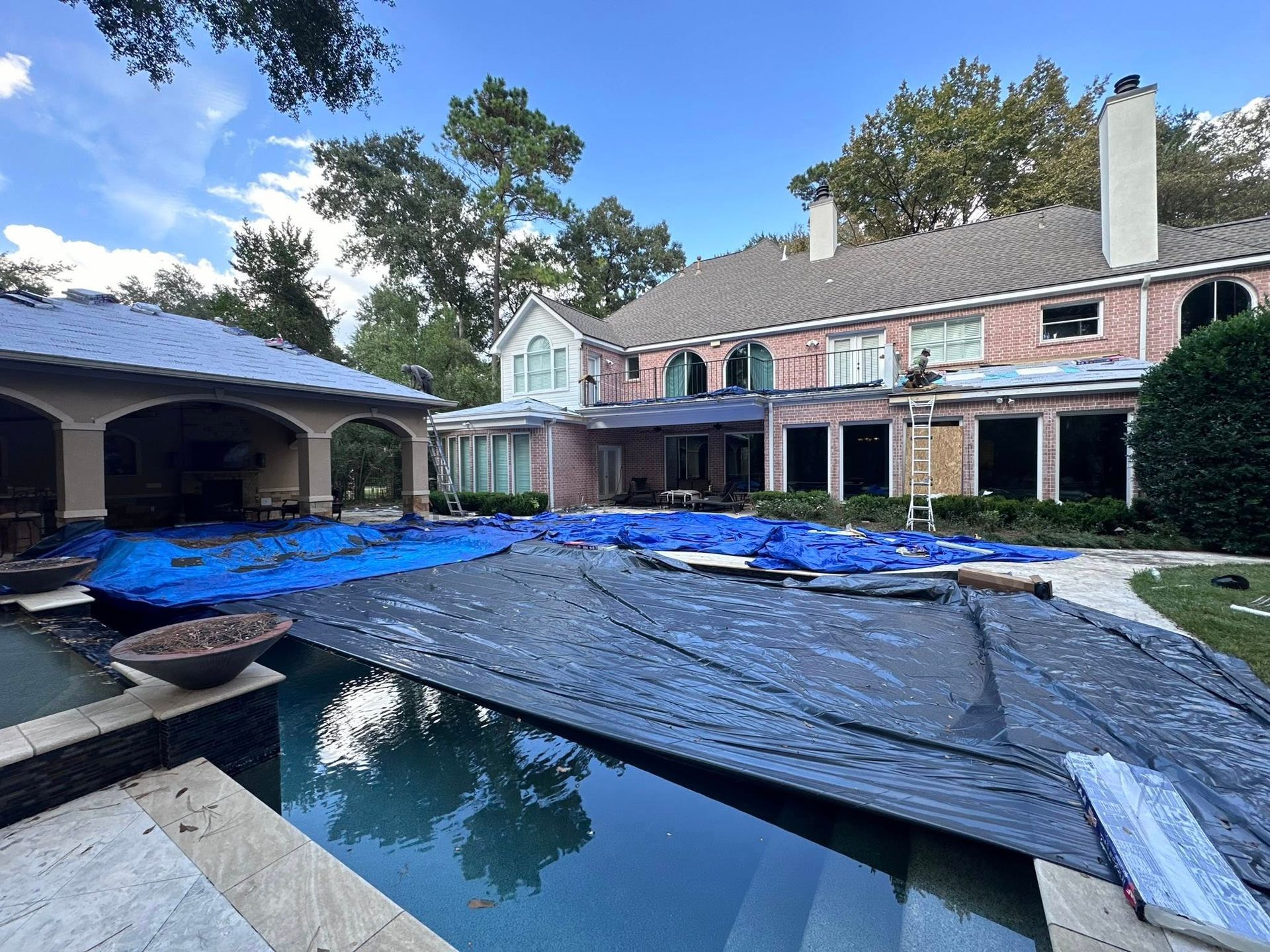 A large brick home with a pool covered by a blue tarp and a covered patio area under a bright, sunny sky.