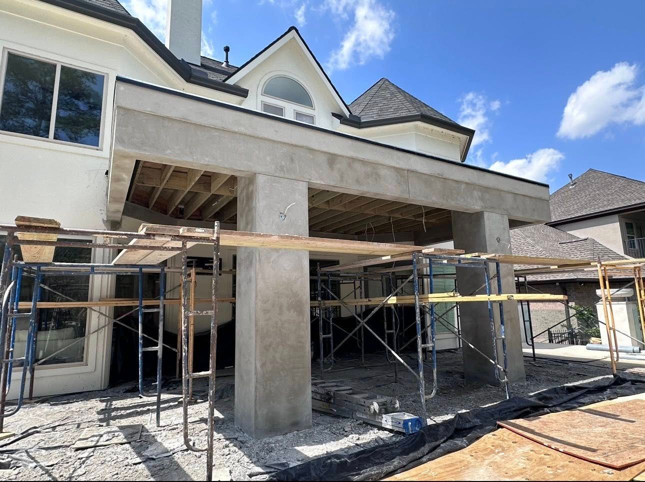 A tan, two-story house under construction with scaffolding around a large, open patio area and exposed wooden ceiling.