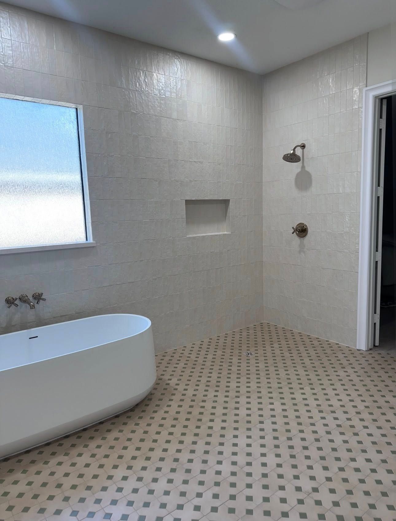 A modern bathroom features a white soaking tub, a tiled walk-in shower, and patterned floor tiles in a light-filled room.