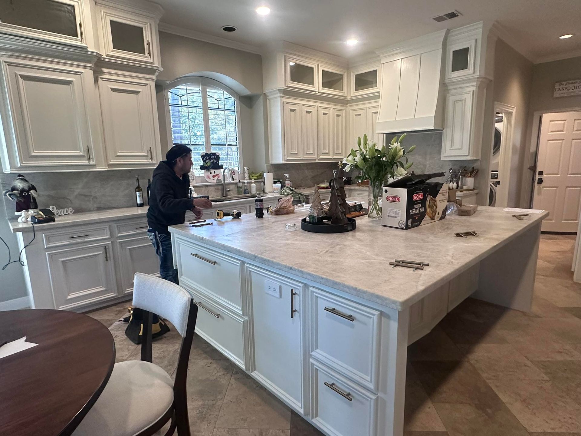 A person stands at a kitchen island in a room with white cabinets, a large stone countertop, and tile flooring.