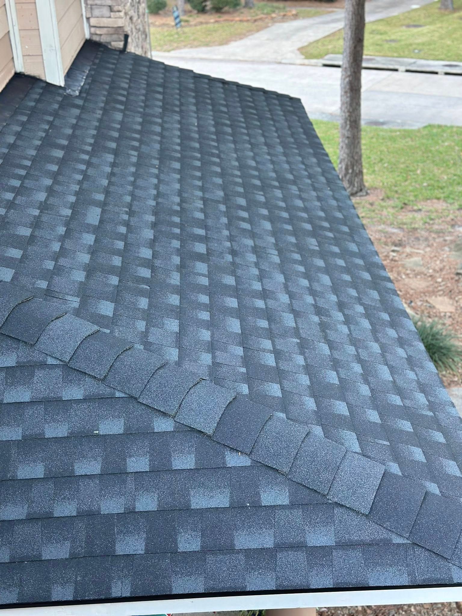 A close-up, high-angle shot of a gray, textured shingle roof sloping downward toward a yard and driveway.