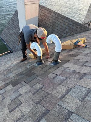 A worker installs two white plumbing vent pipes on a shingled residential roof.