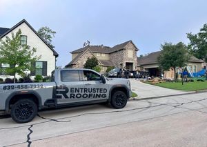 A grey Entrusted Roofing pickup truck parked on a street in front of a house under construction with roofers working.