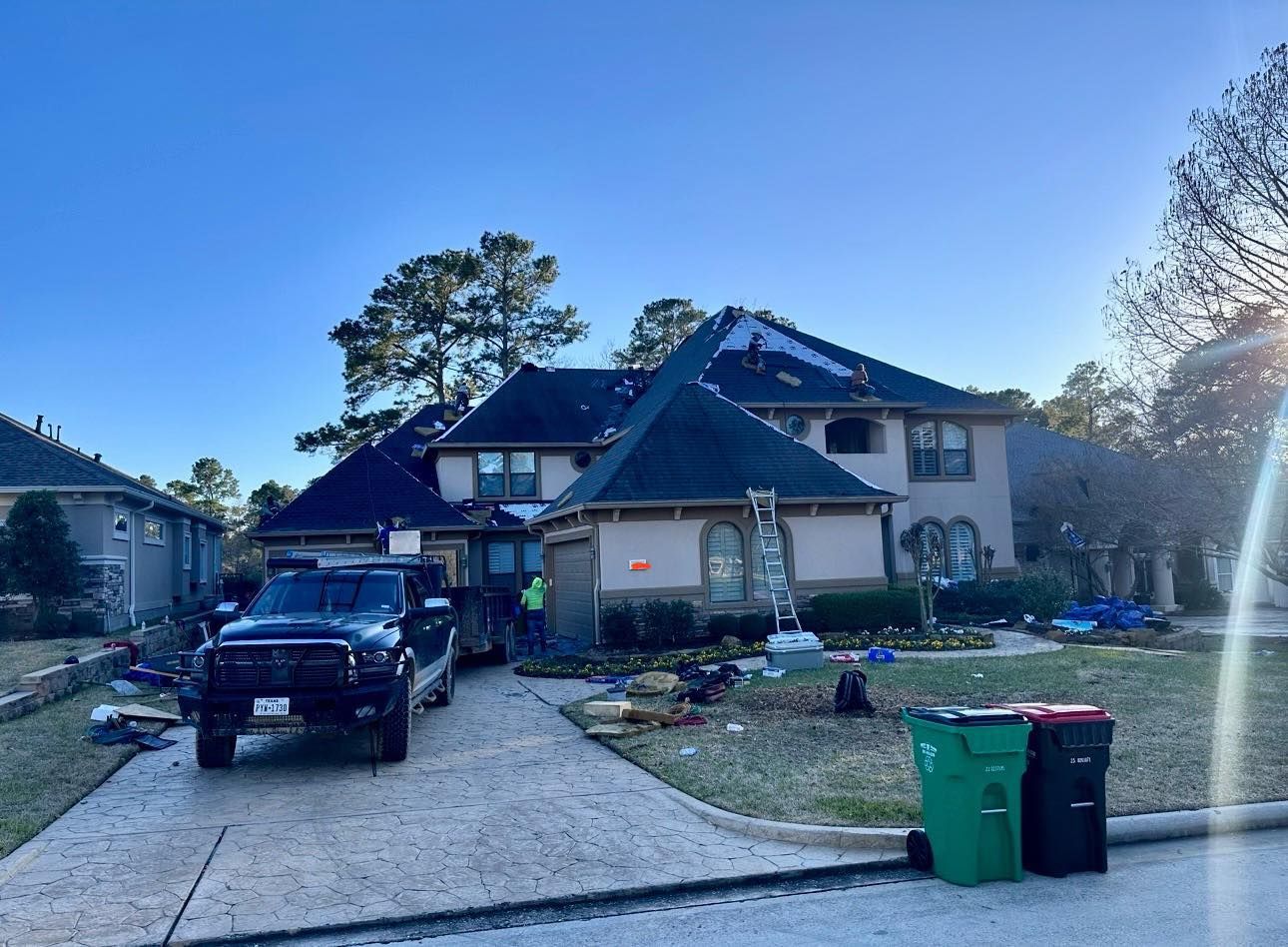 A large multi-story suburban home with a dark roof under renovation, a black truck in the driveway, and waste bins.